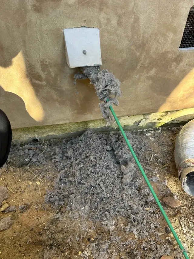 Lint pouring out of a clogged exterior dryer vent on a stucco wall as the vent is cleaned